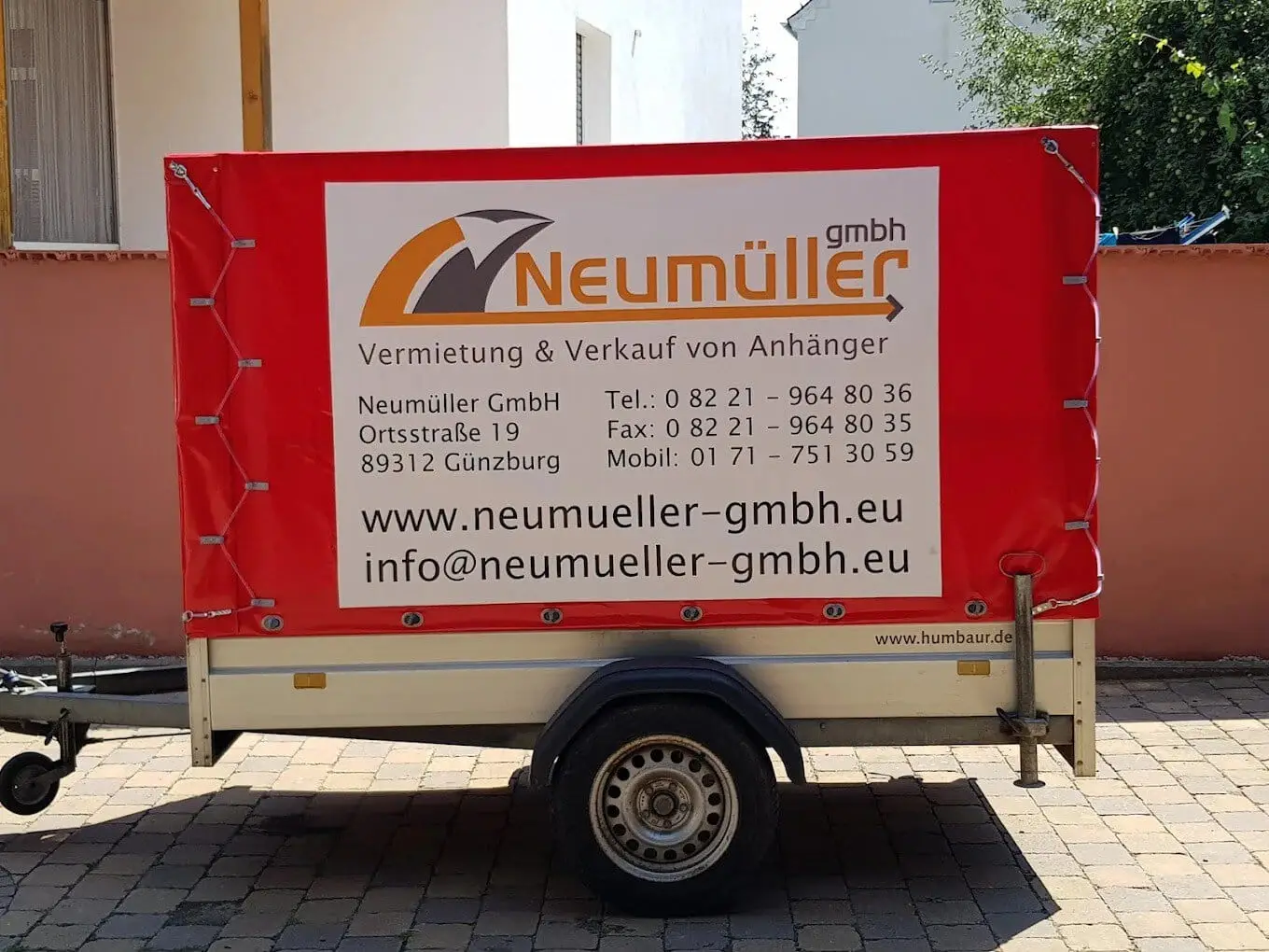 Kleiner Kastenanhänger der Neumüller GmbH mit roter Plane und Firmenbeschriftung